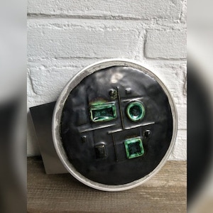 May include: A round, dark gray decorative plate with a silver rim. The plate features geometric shapes and emerald green accents. The plate is resting on a wooden surface in front of a white brick wall.