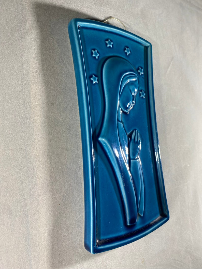 May include: A blue ceramic wall plaque with a raised image of a woman in prayer. The plaque is decorated with white stars.