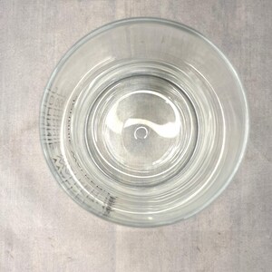 May include: Clear glass tumbler with a circular design, viewed from above. The glass has a wide rim and a slightly textured surface. The glass is empty and sits on a gray surface. The glass has text around the rim.