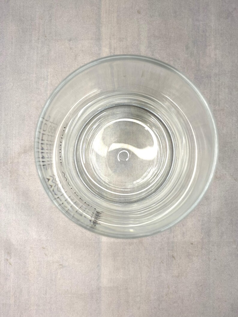 May include: Clear glass tumbler with a circular design, viewed from above. The glass has a wide rim and a slightly textured surface. The glass is empty and sits on a gray surface. The glass has text around the rim.
