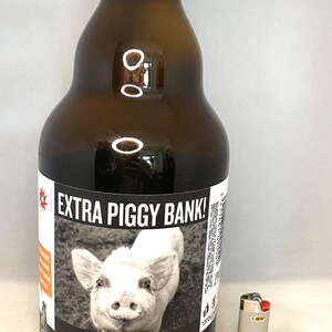 May include: A brown glass bottle with a label that reads "EXTRA PIGGY BANK!" and features a black and white photo of a pig. A silver BIC lighter is next to the bottle. The bottle has a curved shape and a narrow neck.