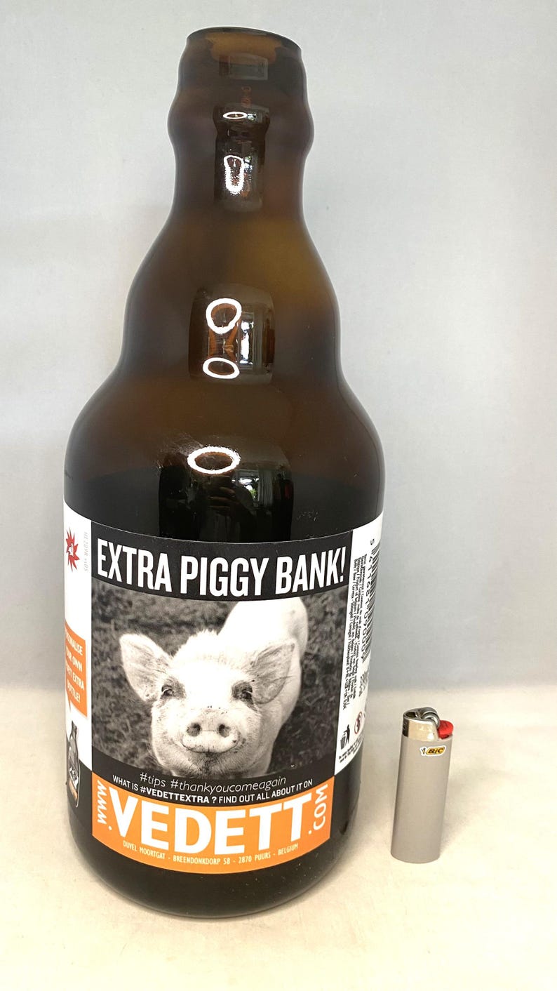 May include: A brown glass bottle with a label that reads "EXTRA PIGGY BANK!" and features a black and white photo of a pig. A silver BIC lighter is next to the bottle. The bottle has a curved shape and a narrow neck.