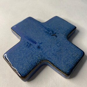 K&ouml;nnte beinhalten: Ein blaues Keramikkreuz mit leicht strukturierter Oberfl&auml;che. Das Kreuz hat eine dunkelblaue Glasur mit einem leichten Metallic-Glanz.