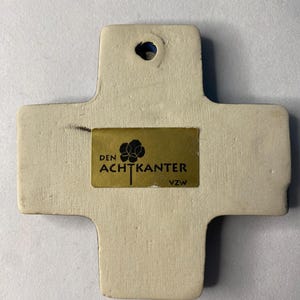 K&ouml;nnte beinhalten: Ein wei&szlig;es Keramikkreuz mit einem goldenen Etikett mit der Aufschrift "DEN ACHTKANTER VZW".