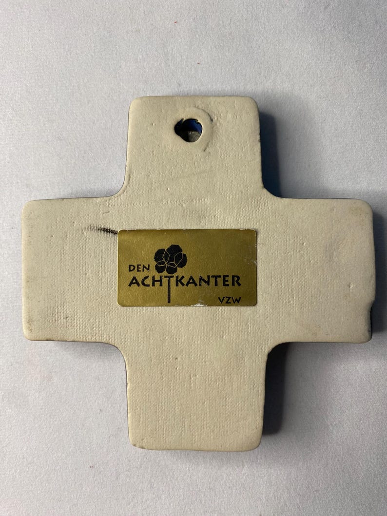 K&ouml;nnte beinhalten: Ein wei&szlig;es Keramikkreuz mit einem goldenen Etikett mit der Aufschrift "DEN ACHTKANTER VZW".