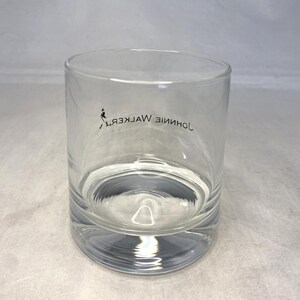 May include: A clear, cylindrical rocks glass with a thick base. The glass features the Johnnie Walker logo and text in black. The glass is partially filled with a clear liquid, likely a beverage.