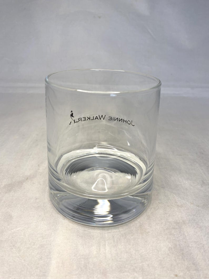 May include: A clear, cylindrical rocks glass with a thick base. The glass features the Johnnie Walker logo and text in black. The glass is partially filled with a clear liquid, likely a beverage.