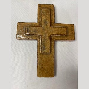 May include: A brown ceramic cross with a raised design. The cross is made of a textured clay and has a rustic look.