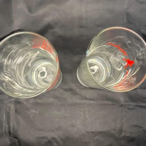 Może przedstawiać: Dwa przezroczyste szklane kieliszki do picia z czerwonym wzorem po bokach. Wz&oacute;r zawiera słowa "Cocktail Classics" i stylizowany obraz koktajlu.