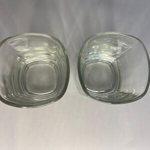 Puede incluir: Dos vasos cuadrados de vidrio transparente, uno ligeramente inclinado hacia un lado, sobre una superficie blanca.