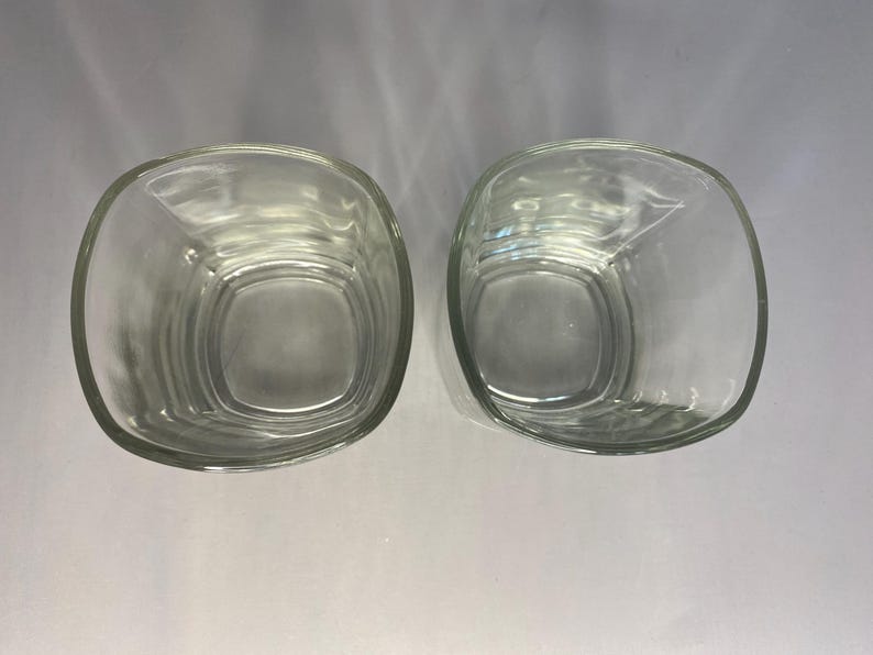 Puede incluir: Dos vasos cuadrados de vidrio transparente, uno ligeramente inclinado hacia un lado, sobre una superficie blanca.