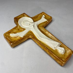 May include: A ceramic cross with a figure in white robes. The cross has a brown and gold finish with a raised relief design. The figure is in a flowing robe with outstretched arms.