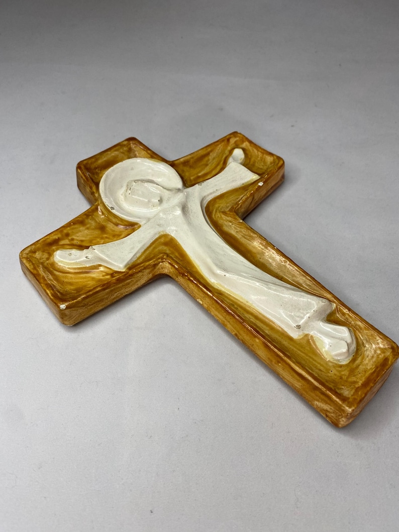 May include: A ceramic cross with a figure in white robes. The cross has a brown and gold finish with a raised relief design. The figure is in a flowing robe with outstretched arms.