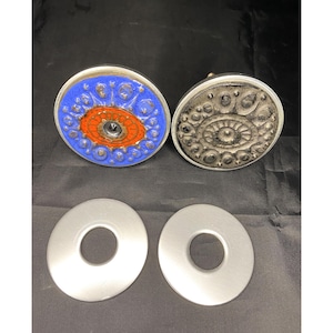 Könnte beinhalten: Zwei dekorative Keramikknöpfe mit komplizierten Designs. Ein Knopf ist blau mit orangefarbenen und schwarzen Akzenten, der andere ist schwarz mit silbernen Akzenten. Zwei silberne Metallringe mit einem Loch in der Mitte.