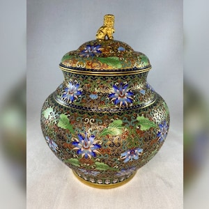 Può includere: Un vaso cloisonné con un coperchio che presenta un ornamento a forma di leone dorato. Il vaso è decorato con intricati motivi floreali in tonalità di blu, verde e oro.
