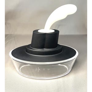 May include: A clear glass bowl with a black lid and a white, curved handle. The bowl has measurement markings. The lid is black with a white rim. The handle is white and curves upwards.