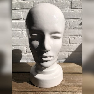 Könnte beinhalten: Ein weißer Keramik-Mannequin-Kopf mit geschlossenem Augenausdruck. Der Kopf ist glatt und hat ein schlichtes, modernes Design.
