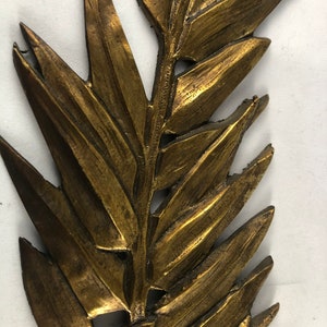 May include: A gold-colored metal wall decor in the shape of a fern leaf. The leaf is detailed and textured, with a realistic appearance.