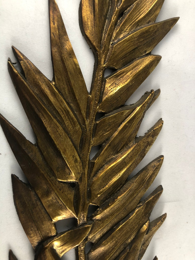 May include: A gold-colored metal wall decor in the shape of a fern leaf. The leaf is detailed and textured, with a realistic appearance.