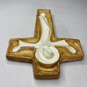 May include: A ceramic cross with a religious theme. The cross is primarily brown with a white central figure. The figure appears to be a person. The cross has a glossy finish and is likely a decorative item.