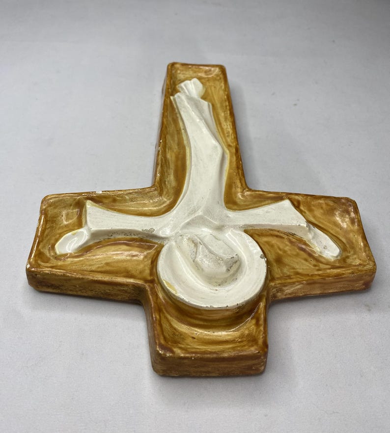 May include: A ceramic cross with a religious theme. The cross is primarily brown with a white central figure. The figure appears to be a person. The cross has a glossy finish and is likely a decorative item.
