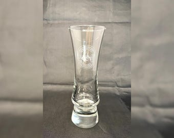 May include: A clear glass shot glass with a wide base and a tall, slender stem. The glass has an etched design of a building with the text "Anderson School of Commerce" around it.