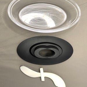 May include: A clear, oval plastic dish is positioned above a black, oval base with a central ring. Beneath, a white and silver utensil with a curved blade and a cross-shaped handle is shown.