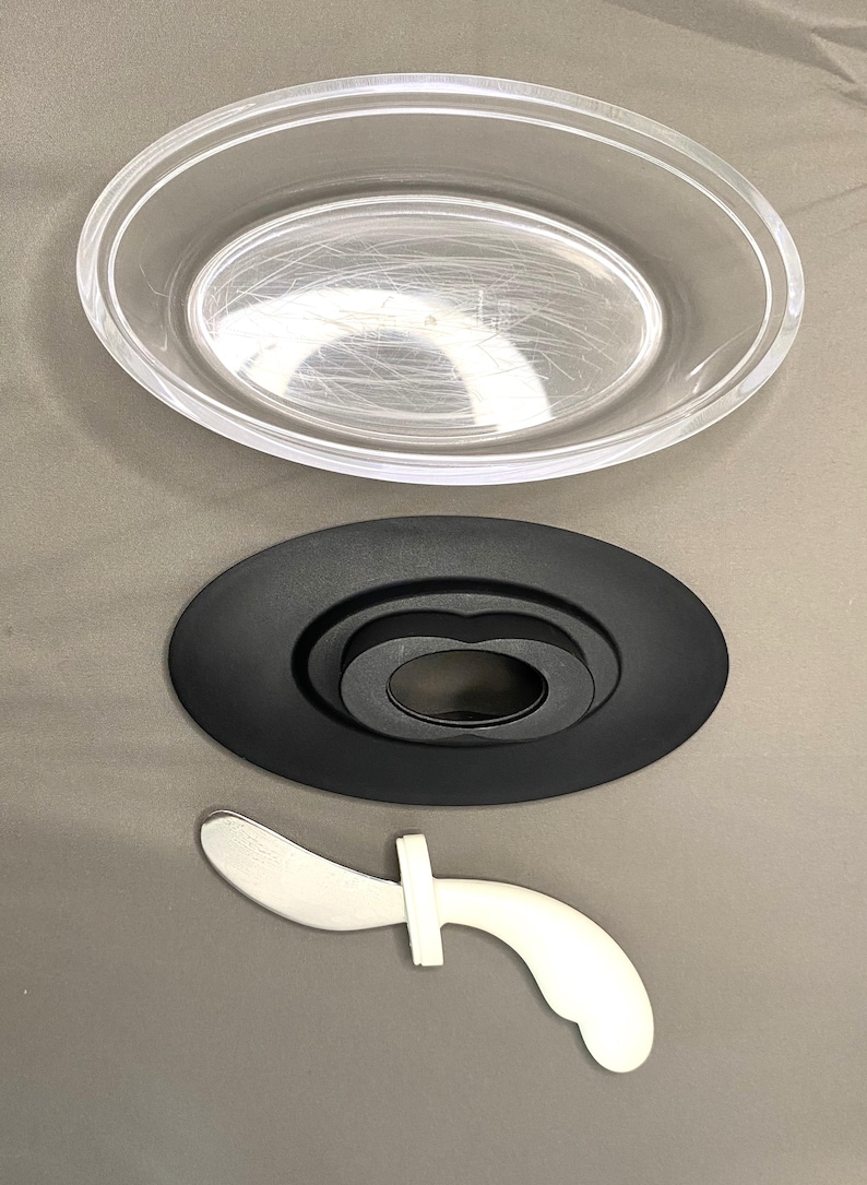 May include: A clear, oval plastic dish is positioned above a black, oval base with a central ring. Beneath, a white and silver utensil with a curved blade and a cross-shaped handle is shown.