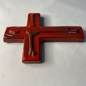 May include: A glossy, red ceramic cross with a detailed depiction of a figure. The cross has a rectangular base and a smooth, reflective surface. The figure's form is visible, with dark brown accents enhancing the details.