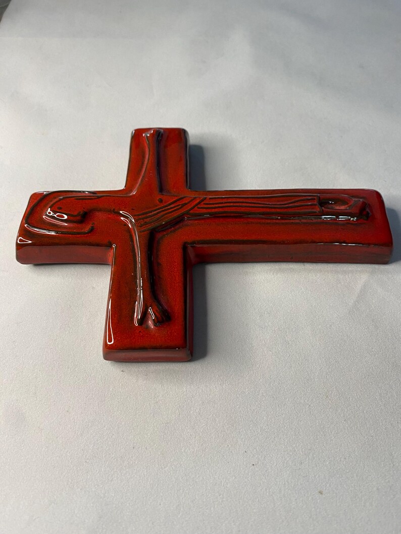 May include: A glossy, red ceramic cross with a detailed depiction of a figure. The cross has a rectangular base and a smooth, reflective surface. The figure's form is visible, with dark brown accents enhancing the details.