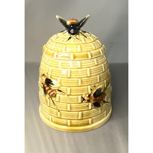 Könnte beinhalten: Gelber Keramik-Honigtopf in Form eines Bienenstocks, mit einem Deckel, der eine Biene zeigt. Der Topf hat ein strukturiertes Ziegelmuster und ist mit zwei gemalten Bienenillustrationen verziert.