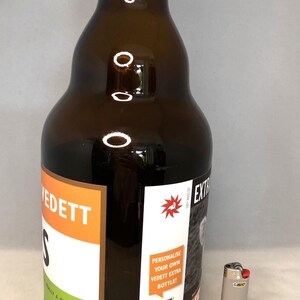 May include: A brown glass beer bottle with a white and orange label, and a silver Bic lighter. The bottle has the text "VEDETT" and "WALLY A PALE ALE" on the label. The bottle contains 330ml of beer.