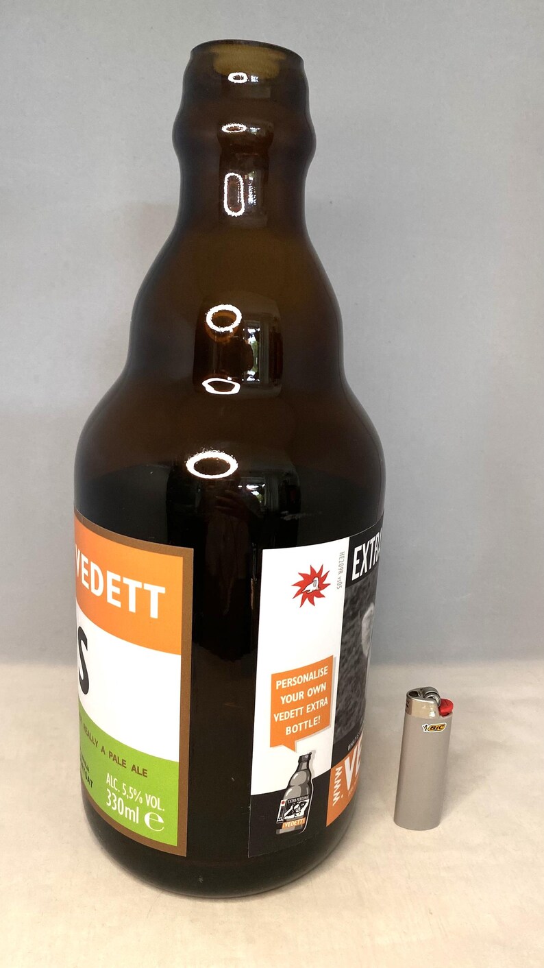 May include: A brown glass beer bottle with a white and orange label, and a silver Bic lighter. The bottle has the text "VEDETT" and "WALLY A PALE ALE" on the label. The bottle contains 330ml of beer.