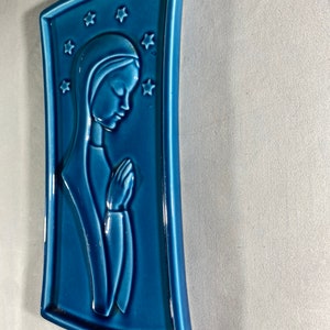May include: A blue ceramic wall plaque with a raised image of a woman in prayer. The woman is wearing a blue robe and has her hands clasped in front of her. The plaque is decorated with stars.