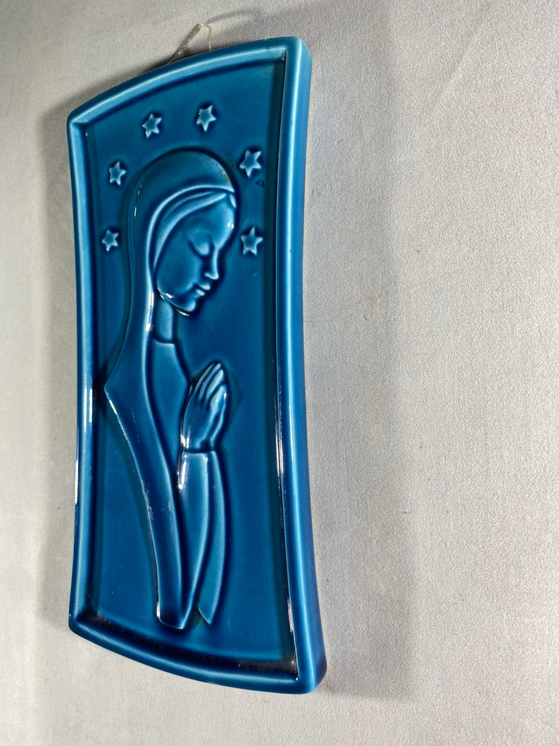 May include: A blue ceramic wall plaque with a raised image of a woman in prayer. The woman is wearing a blue robe and has her hands clasped in front of her. The plaque is decorated with stars.