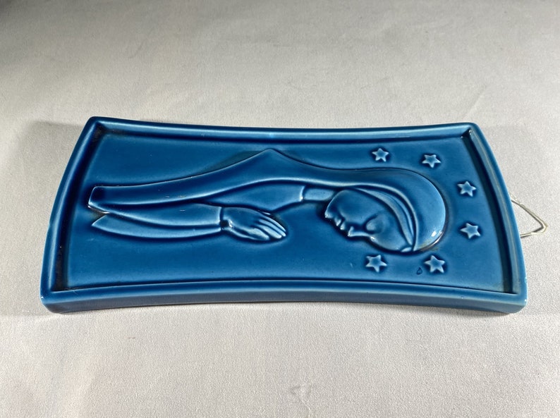 May include: A blue ceramic wall plaque with a raised image of a woman in a blue robe with stars around her head.
