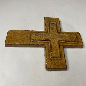 May include: A brown ceramic cross with a raised design. The cross is made of a textured clay and has a rustic look.