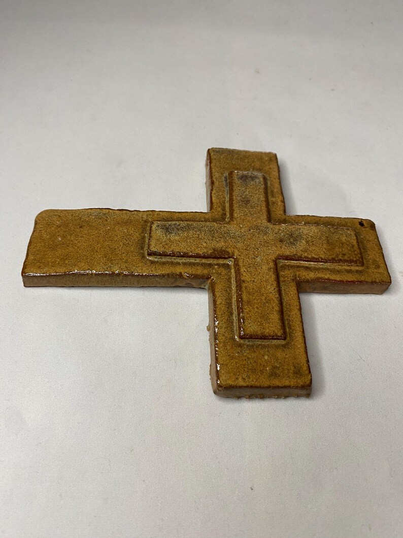 May include: A brown ceramic cross with a raised design. The cross is made of a textured clay and has a rustic look.