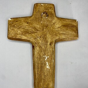 May include: A handcrafted, textured cross with a warm, golden-brown finish. The religious symbol features a loop at the top for hanging. The cross has a rustic, aged appearance, with white accents along the edges.