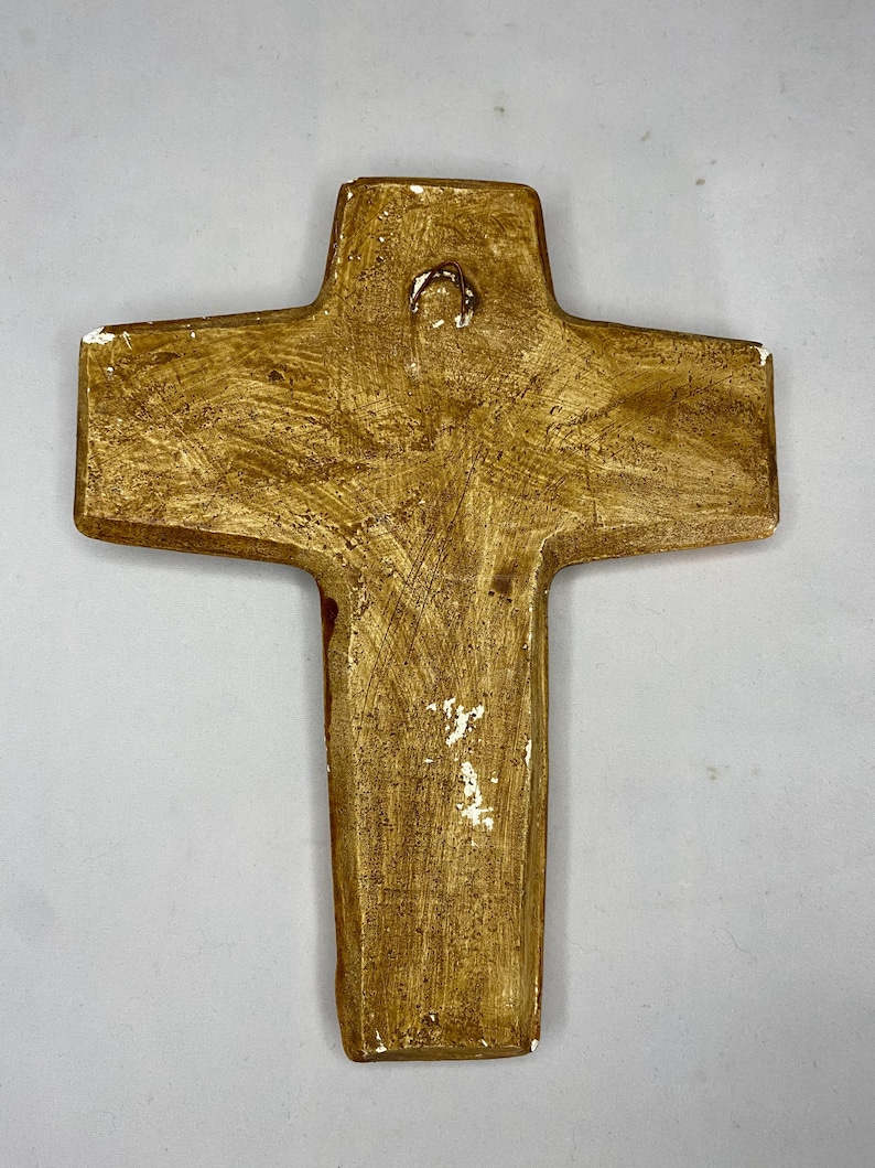 May include: A handcrafted, textured cross with a warm, golden-brown finish. The religious symbol features a loop at the top for hanging. The cross has a rustic, aged appearance, with white accents along the edges.