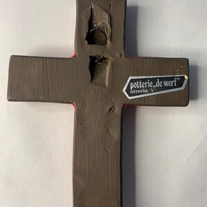 May include: A brown ceramic cross with a red border. The back of the cross has a metal wire for hanging and a sticker that reads "potterie, de werf" and "utrecht". The cross is a religious item.