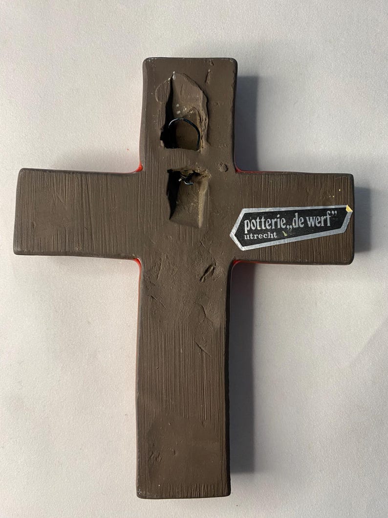 May include: A brown ceramic cross with a red border. The back of the cross has a metal wire for hanging and a sticker that reads "potterie, de werf" and "utrecht". The cross is a religious item.