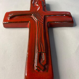 May include: A red ceramic cross with a glossy finish. The cross features a relief of a figure with outstretched arms. The figure's form is detailed with dark brown lines, and the cross has a rectangular shape.