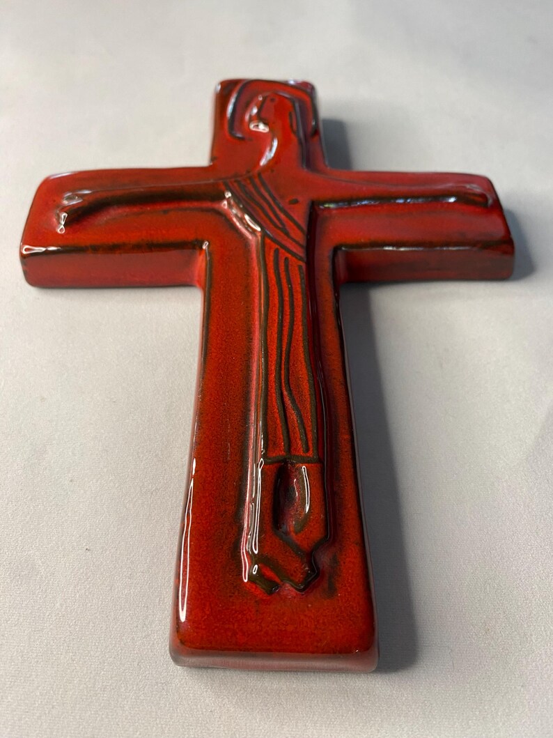 May include: A red ceramic cross with a glossy finish. The cross features a relief of a figure with outstretched arms. The figure's form is detailed with dark brown lines, and the cross has a rectangular shape.
