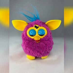 May include: A pink and yellow Furby toy with blue hair and large, blue eyes. The toy has a yellow beak and a red mouth.