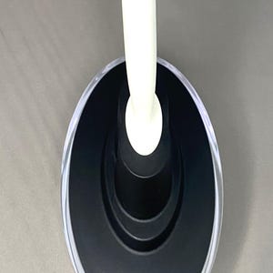 May include: An oval-shaped, black and clear soap dish with a white handle. The dish has a clear, transparent rim and a black interior. The white handle is curved and extends upwards from the centre of the dish.