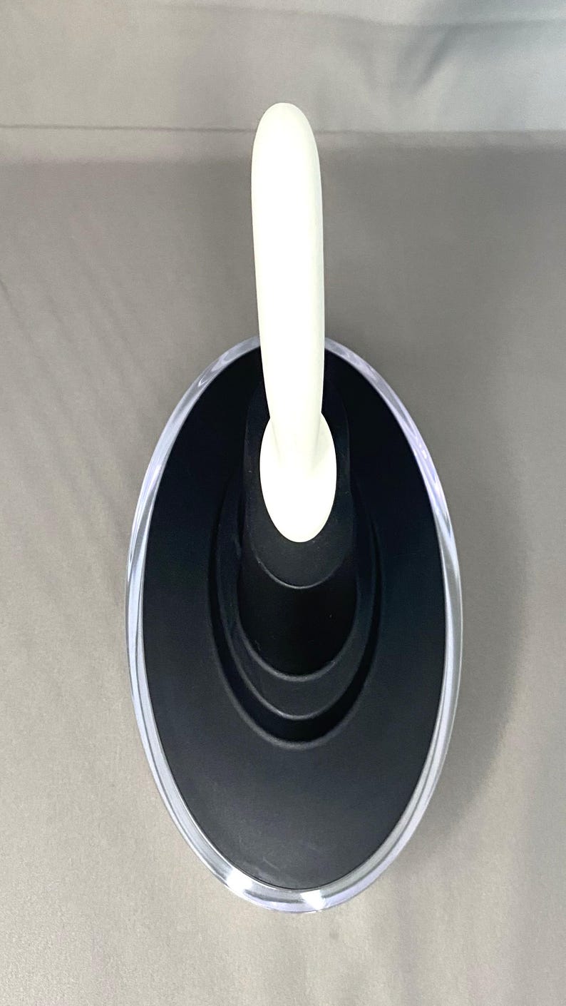May include: An oval-shaped, black and clear soap dish with a white handle. The dish has a clear, transparent rim and a black interior. The white handle is curved and extends upwards from the centre of the dish.
