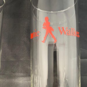 Może przedstawiać: Dwa przezroczyste szklanki z czerwonym logo Johnnie Walker i sylwetką idącego mężczyzny.