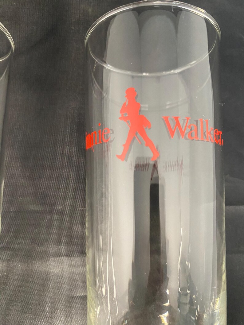 Może przedstawiać: Dwa przezroczyste szklanki z czerwonym logo Johnnie Walker i sylwetką idącego mężczyzny.