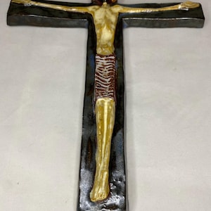 Może przedstawiać: Ceramiczny krzyż ścienny z ciemnoniebieską glazurą i beżową figurą Jezusa Chrystusa. Figura jest przedstawiona w brązowym przepasaniu i z wyciągniętymi ramionami.